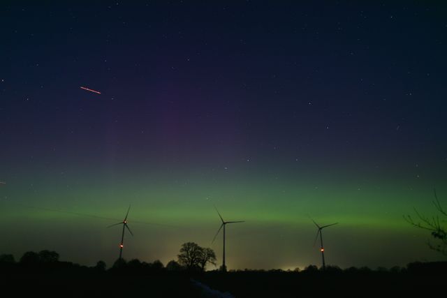 Aurora boreal tenue cerca del horizonte sobre un paisaje nocturno con aerogeneradores