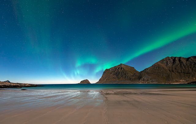 Aurora boreal sobre una playa y montañas bajo un cielo estrellado